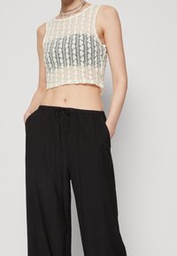 Krajkový crop top v krémové barvě s pruhovaným vzorem a scalloped lemem, kombinovaný s volnými černými kalhotami s šňůrkou.