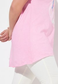 Camiseta de algodón rosa con un dobladillo curvado y un poco más larga por detrás, con un diseño gráfico sencillo. Combinada con leggings blancos.