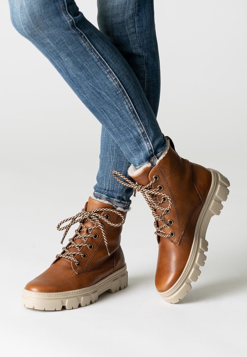 Personne portant un jean skinny bleu et des bottes en cuir marron à lacets avec des semelles épaisses beige, debout sur un sol blanc.
