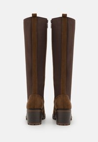 Bottes hautes marron avec talons carrés et fermeture éclair arrière, présentant des accents en daim et une semelle texturée, vues de dos sur fond blanc.
