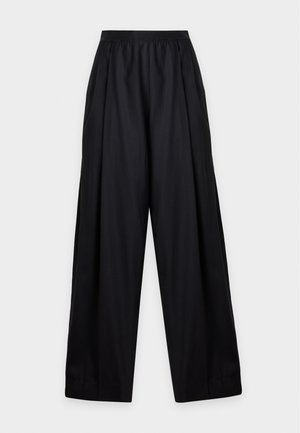Femme aux longs cheveux noirs portant un haut crème sans manches et un pantalon large noir ample, debout avec les mains sur les hanches.