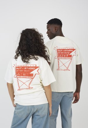 Dwie osoby ubrane w kremowe koszulki z czerwonym napisem "OUT of STUDIO" na plecach, w połączeniu z jasnoniebieskimi dżinsami, na białym tle.