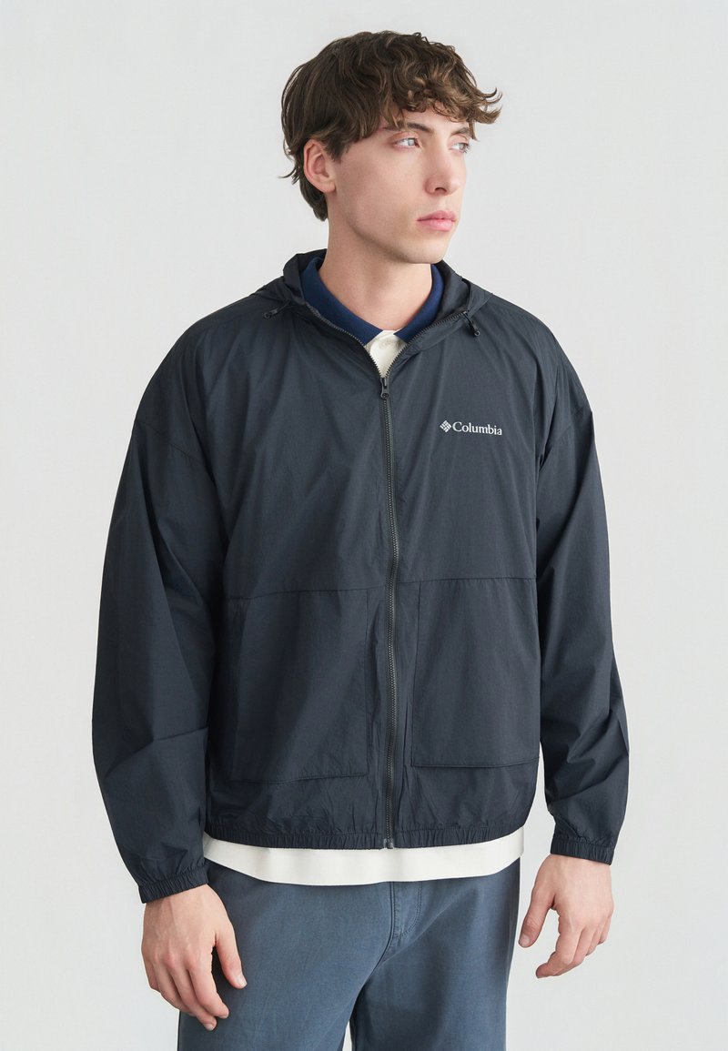 Νεαρός άνδρας με μαύρο φούτερ Columbia με φερμουάρ και κουκούλα πάνω από λευκό και ναυτικό πουκάμισο, στέκεται με χαλαρά τα χέρια, κοιτάζοντας δεξιά σε απλό φόντο.