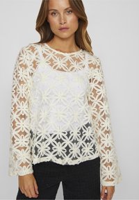 Haut à manches longues en mesh blanc avec broderies florales, superposé sur un débardeur blanc. Présente un col rond et des manches évasées, avec un motif texturé.