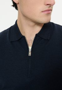 Primo piano di un uomo che indossa un maglione blu navy con una mezza cerniera argentata, colletto piegato e peli del petto chiari visibili attraverso l'apertura della cerniera.