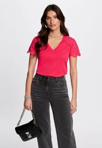 Camiseta de cuello en V rosa con mangas de encaje con ojales en forma de concha, combinada con vaqueros de mezclilla negros de cintura alta y un bolso negro acolchado.