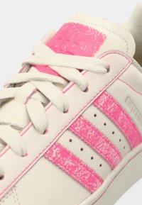 Balti odiniai sportbačiai su rausvomis tekstūruotomis detalėmis, kuriuose yra trys rausvos juostos, balti raišteliai ir prekės ženklo kalbos žymė.