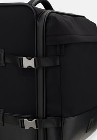 Czarna torba podróżna na plecy wykonana z nylonu i skóry, charakteryzująca się prostokątnym kształtem, płaskim dnem, bocznymi paskami z klamrami i małą naszywką z logo.