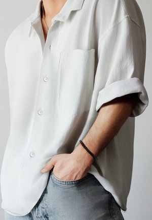 Camisa gris claro de manga corta con botones y un bolsillo delantero, hecha de una tela texturizada. Llevada sobre unos vaqueros azules, con una pulsera negra.