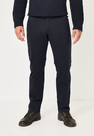Mann trägt dunkelblaue Hose, schwarze Schnürstiefel aus Leder und einen dunklen Strickpullover, steht vor einem schlichten hellen Hintergrund.
