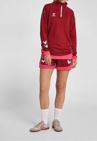 Sudadera deportiva roja con cremallera, logo blanco y detalles en rosa, combinada con pantalones cortos rojos con ribetes rosas y detalles en chevron blancos. Zapatillas deportivas grises.