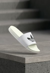 Hvit slip-in sandal med svart Adidas-logo og tre striper, med glatt plastoverdel, flat såle og minimalistisk design.
