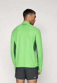 Αθλητική μπλούζα με μακριά μανίκια σε έντονο πράσινο χρώμα, με σκουροπράσινες πλευρικές πινελιές και ψηλό γιακά. Το υλικό φαίνεται υφασμάτινο και εφαρμοστό.