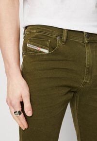 Olive green denim jeans with a slim fit, featuring contrast stitching, a small pocket label, and a front coin pocket.