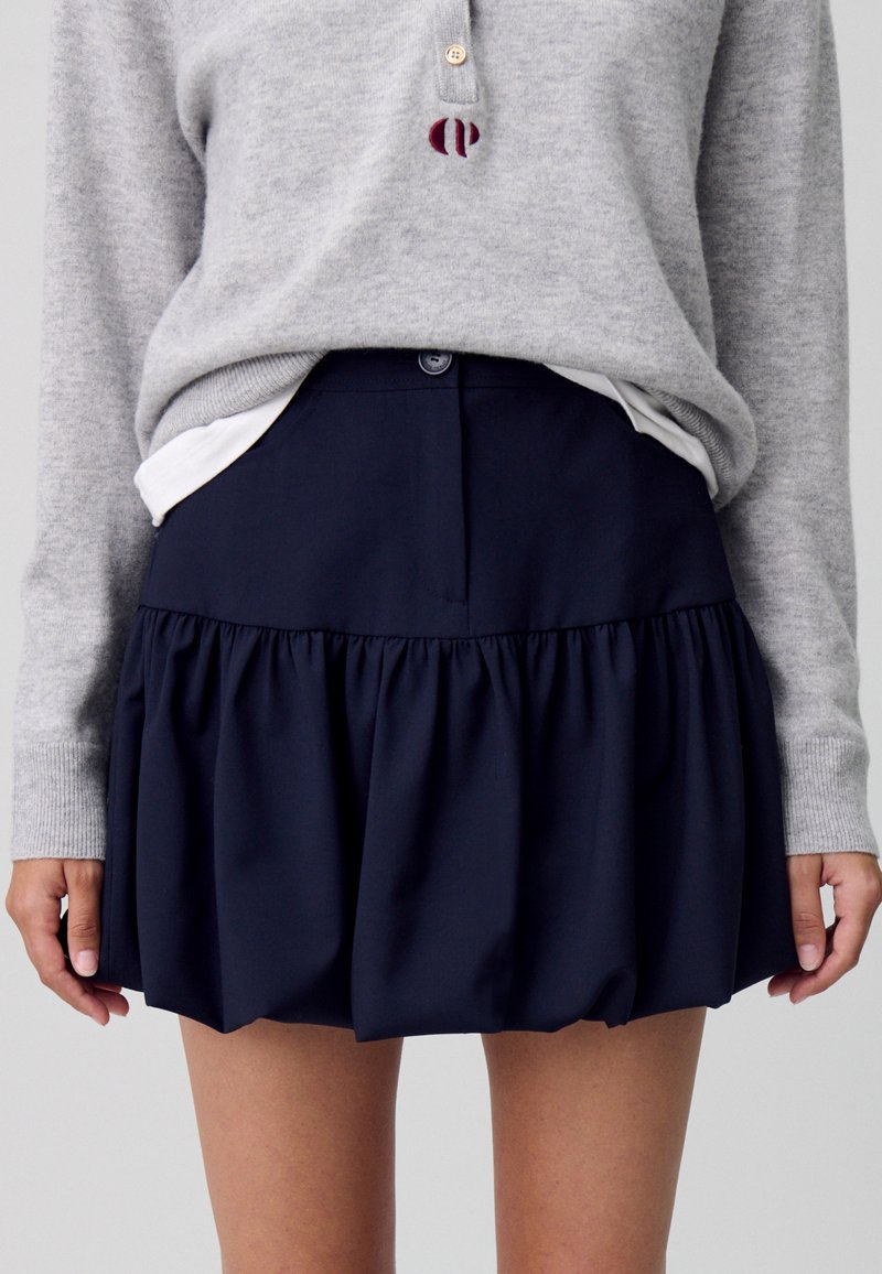 Jupe plissée bleu marine avec une taille ajustée et une fermeture à boutons, confectionnée dans un tissu lisse ; associée à un pull en maille gris clair.