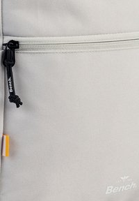 Grijze stoffen rugzak met een gladde textuur, voorzien van een ritsvak, zwarte ritsgreep en een klein oranje label. Merklogo zichtbaar.