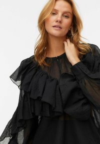 Černá blůza se zdobením v podobě volánů, lehký průhledný materiál, kulatý výstřih a manžety na knoflíky. Texturovaný, splývavý design.