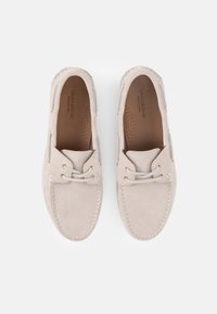 Ζευγάρι μπεζ καστόρινων casual μοκασίνια με χρυσά μεταλλικά τρύπες και ανοιχτόχρωνα κορδόνια, εμφανιζόμενα από πάνω σε λευκό φόντο.