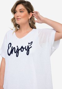 Top de malla blanca con un corte holgado, escote en V, mangas cortas y la palabra "Enjoy" bordada en azul marino con un detalle de corazón.