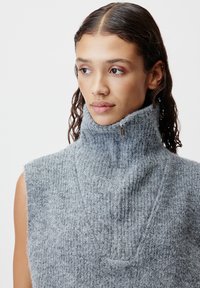 Junge Frau mit nassen, nach hinten gekämmten Haaren, die einen ärmellosen, hochgeschlossenen grauen Strickpullover mit einem Reißverschluss am Kragen trägt.