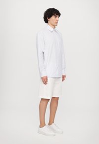 Camicia a righe azzurra chiara, maniche lunghe, con bottoni sul davanti, abbinata a pantaloni corti bianchi. Il modello indossa scarpe da ginnastica bianche e si trova davanti a uno sfondo semplice.