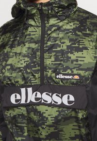 Grøn camouflagejakke med en hætte med lynlås, sorte accenter og et fremtrædende hvidt logo på brystet. Letvægts, struktureret stof.