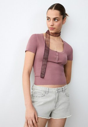 Jeune femme portant un haut court côtelé mauve à manches courtes avec des boutons, une écharpe transparente attachée autour du cou, et un short en denim gris clair, debout contre un mur blanc.