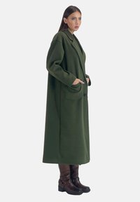 Lång grön ullkappa med kavajkrage, sidfickor och knäppning med en knapp, kombinerad med bruna knähöga stövlar med metallnitar.