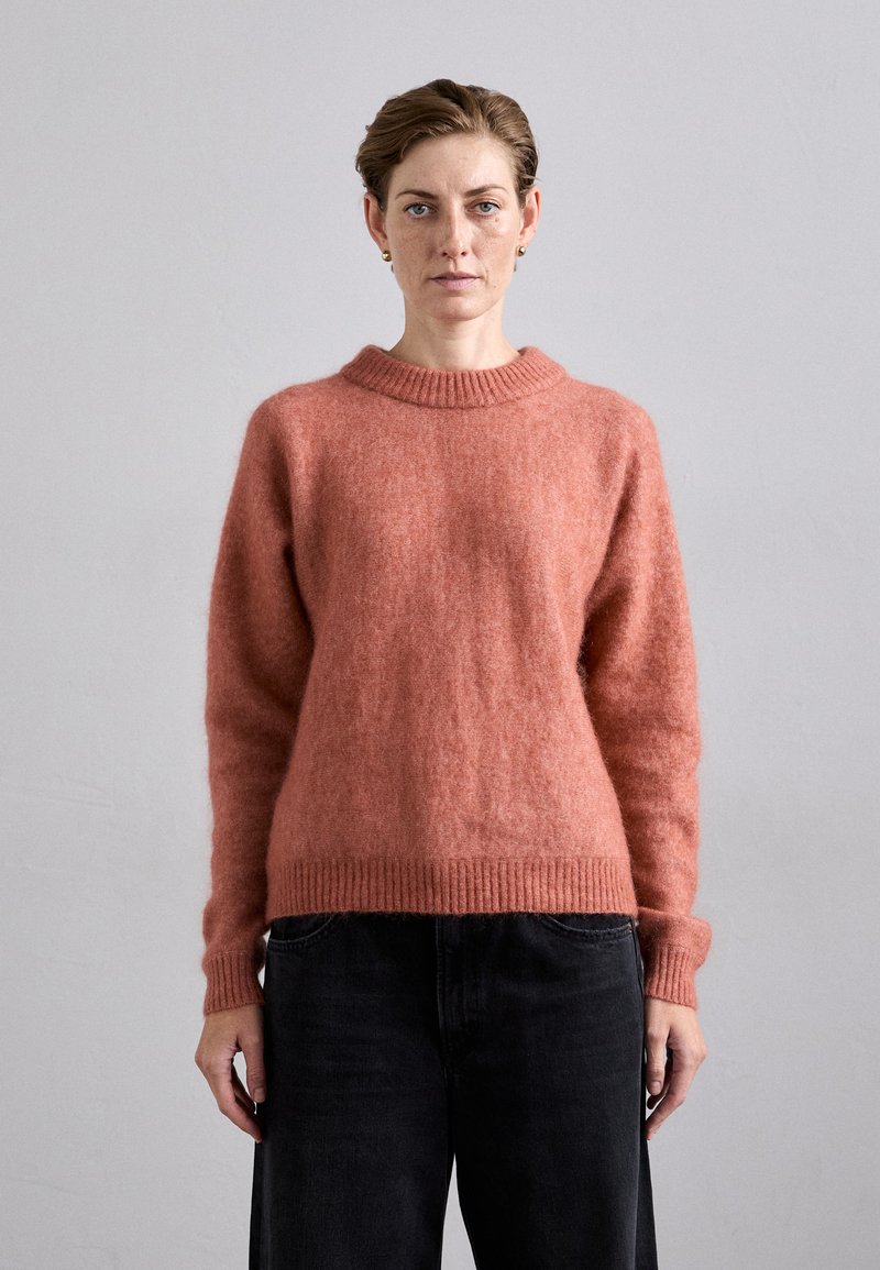 Oversized, round-neck sweater in soft, textured coral wool. Features ribbed cuffs and hem, paired with dark wide-leg jeans.
