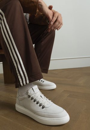 White leather sneakers with grey accents, round toes, and metal eyelets, paired with brown track pants featuring white stripes.