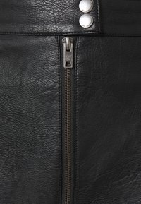 Detailní záběr na černou koženou bundu s kovovým zipem a dvěma stříbrnými patkovými knoflíky, na kterých je vyryto "Pepe Jeans London."