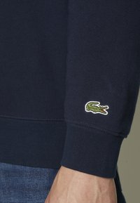 Tamsiai mėlynas megztinis su ilgomis rankovėmis, raukšlėtomis rankogaliais ir žaliu krokodilo logotipu ant kairės rankovės. Pagamintas iš medvilnės medžiagos su lygiu paviršiumi.