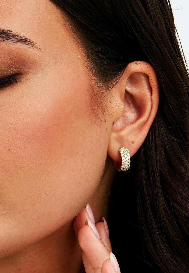 Goldene Creolen mit strukturierter Oberfläche, akzentuiert durch kleine klare Strasssteine, am Ohr vor neutralem Hintergrund gezeigt.