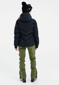 Zwarte pufferjack met chevronstiksel, groene skibroek en zwarte laarzen. Gehaakte zwarte muts met pompon en een skibril op het voorhoofd.