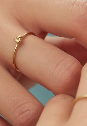 Goldring mit Knotendetail, glatter Oberfläche und dünnem Band. Zwei Finger zeigen den Ring vor einem sanften blauen Hintergrund.