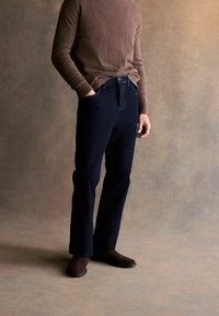 Jeans din denim albastru marin cu picioare drepte, asociate cu un pulover din tricot maro. Modelul stă pe un fundal bej texturat.