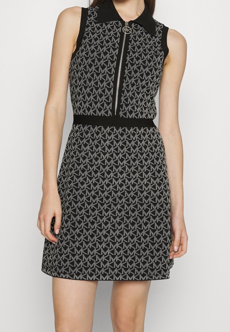 Robe sans manches noire et grise à motifs avec fermeture éclair à l'avant, col noir et ceinture à la taille, portée par une personne debout, les bras détendus.