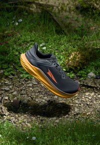 Chaussure de course noire avec une tige texturée, accents orange et semelle jaune rembourrée, posée sur une surface naturelle en pierre.