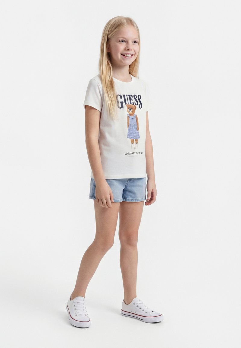 Fille souriante aux longs cheveux blonds portant un t-shirt blanc avec un motif d'ourson, un short en jean et des baskets blanches, debout sur un fond blanc.