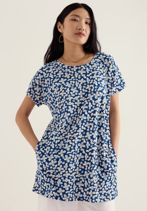 Woman wearing a blue short-sleeve dress with white floral print, gold chain necklace, and gold hoop earrings, hands in pockets.