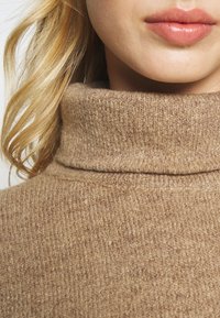 Beige coltrui met een gestructureerd breipatroon. De stof lijkt zacht en warm, met een gevouwen halslijn en een lichtgewicht ontwerp.