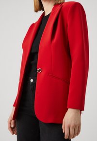 Blazer rouge en tissu lisse, avec un seul bouton en métal, devant ouvert, et deux poches latérales avec des détails passepoilés. Haut noir en dessous.