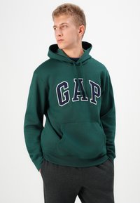 Green hoodie made of cotton blend, featuring a large white and navy "GAP" logo, a front kangaroo pocket, and a drawstring hood.