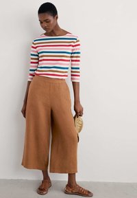 Gestreiftes Langarmshirt mit rosa, blauen und braunen Linien; braune weit geschnittene Leinenhose; braune Sandalen; hält eine Strohtasche.
