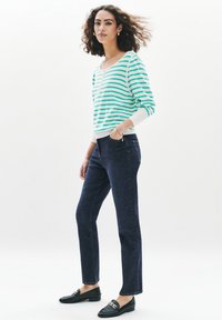 Pull en maille rayé vert et blanc, pantalon en denim droit sombre, et mocassins noirs avec détail en chaîne dorée. Texture lisse, coupe décontractée.