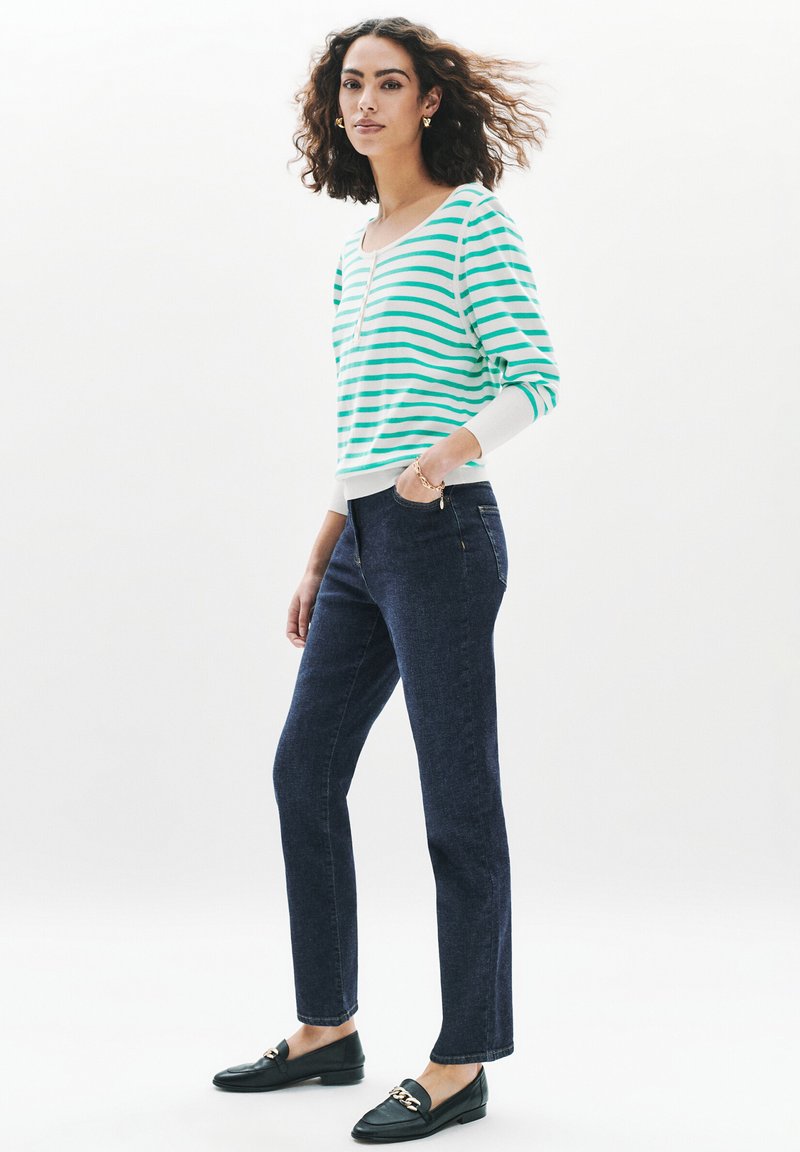 Pull en maille rayé vert et blanc, pantalon en denim droit sombre, et mocassins noirs avec détail en chaîne dorée. Texture lisse, coupe décontractée.
