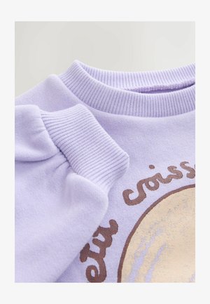 Lillasinine dressipluus soonikkoelise kaeluse ja varrukaservadega, millel on ümmargune graafika tekstiga "croissant" ning pruunide ja kuldsete tekstuursete detailidega.