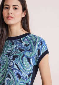 Tee de manga corta con un patrón de paisley azul y verde sobre un fondo marino, con cuello y puños de canalé en color marino sólido.