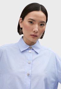 Camicia a righe blu chiaro con colletto button-down, caratterizzata da decorazioni argentate e una vestibilità morbida. I dettagli notevoli includono bottoni arrotondati.