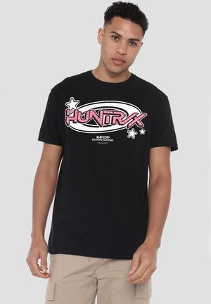 Young man wearing black T-shirt with pink "HUNTR X" text and stars graphic, paired with beige cargo shorts, standing against plain background.